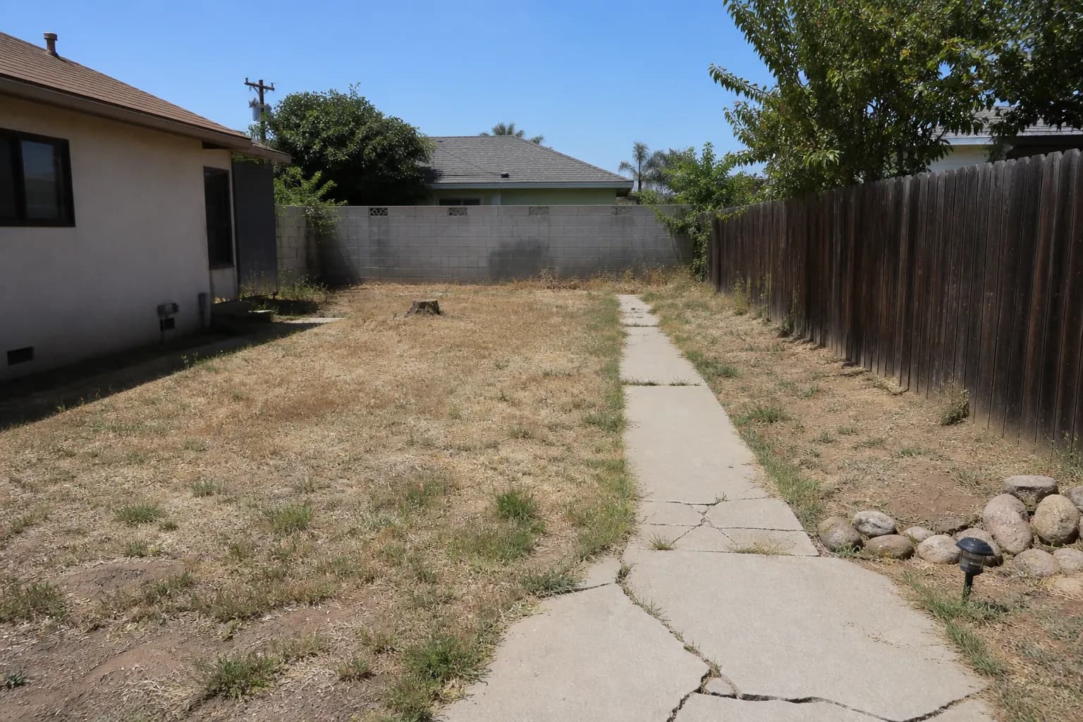 A neglected backyard with a patchy dry lawn, a cracked concrete path and a bare fence in flat light.