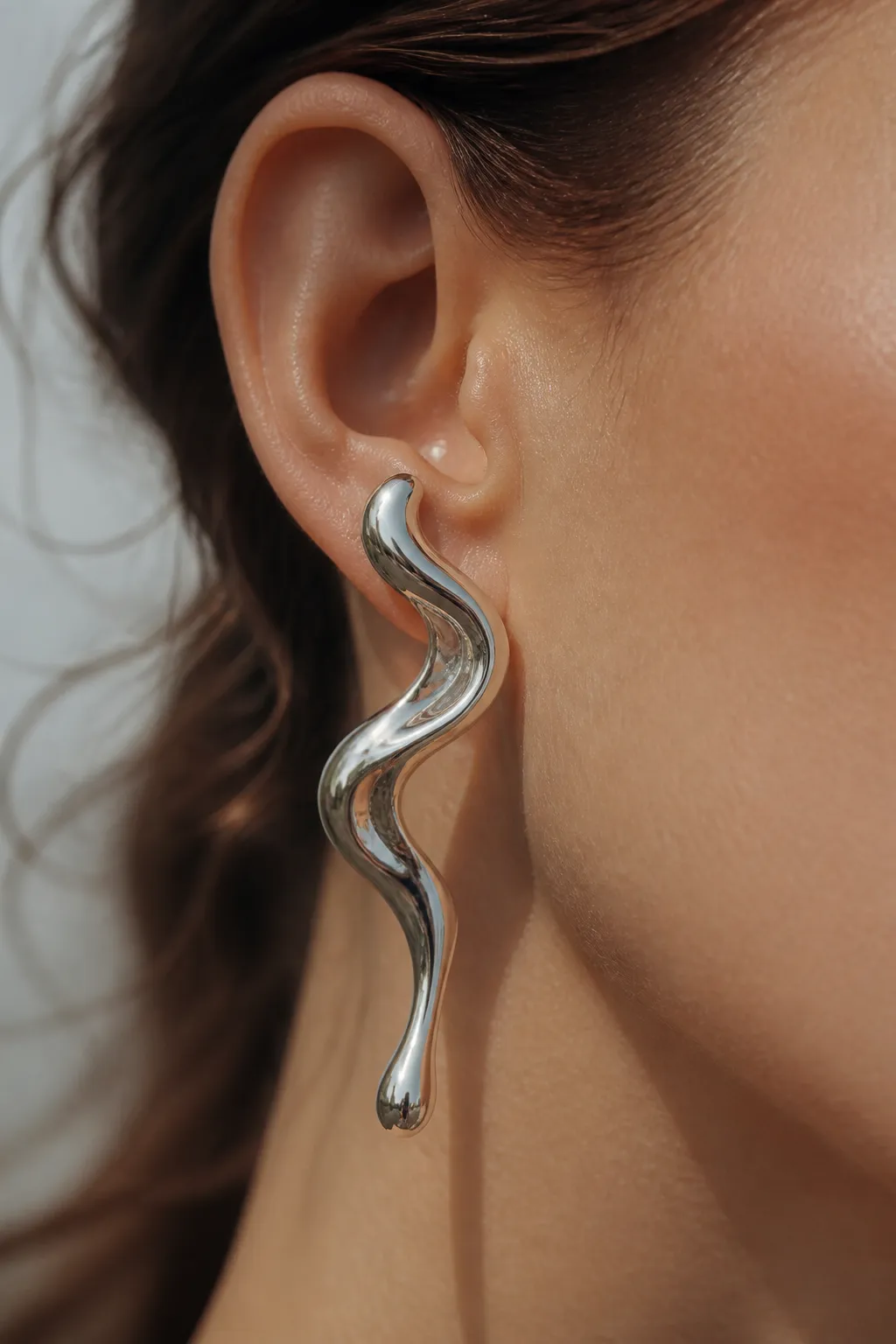 An ear in profile with a sculptural silver earring flowing like a ribbon of liquid metal toward the jaw.