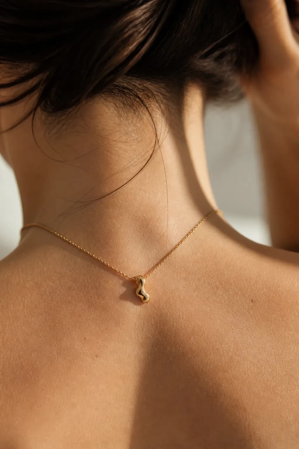 The nape of a neck with the hair lifted, wearing a fine gold chain with a small fluid pendant.