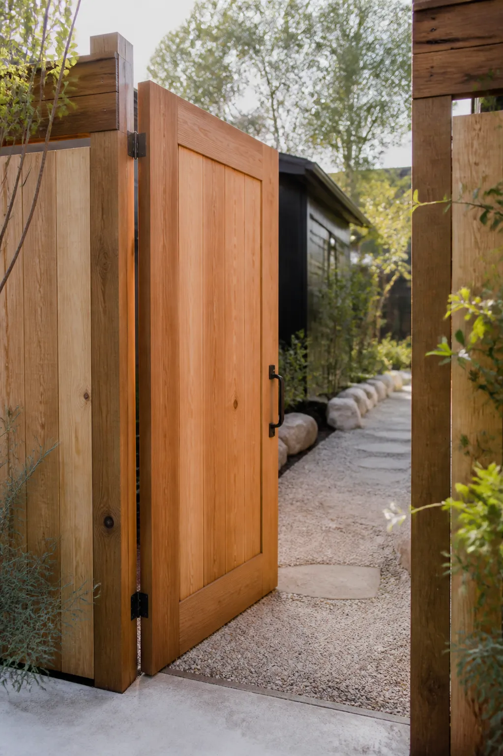 A new cedar garden gate with simple steel hardware standing half open onto a gravel path.