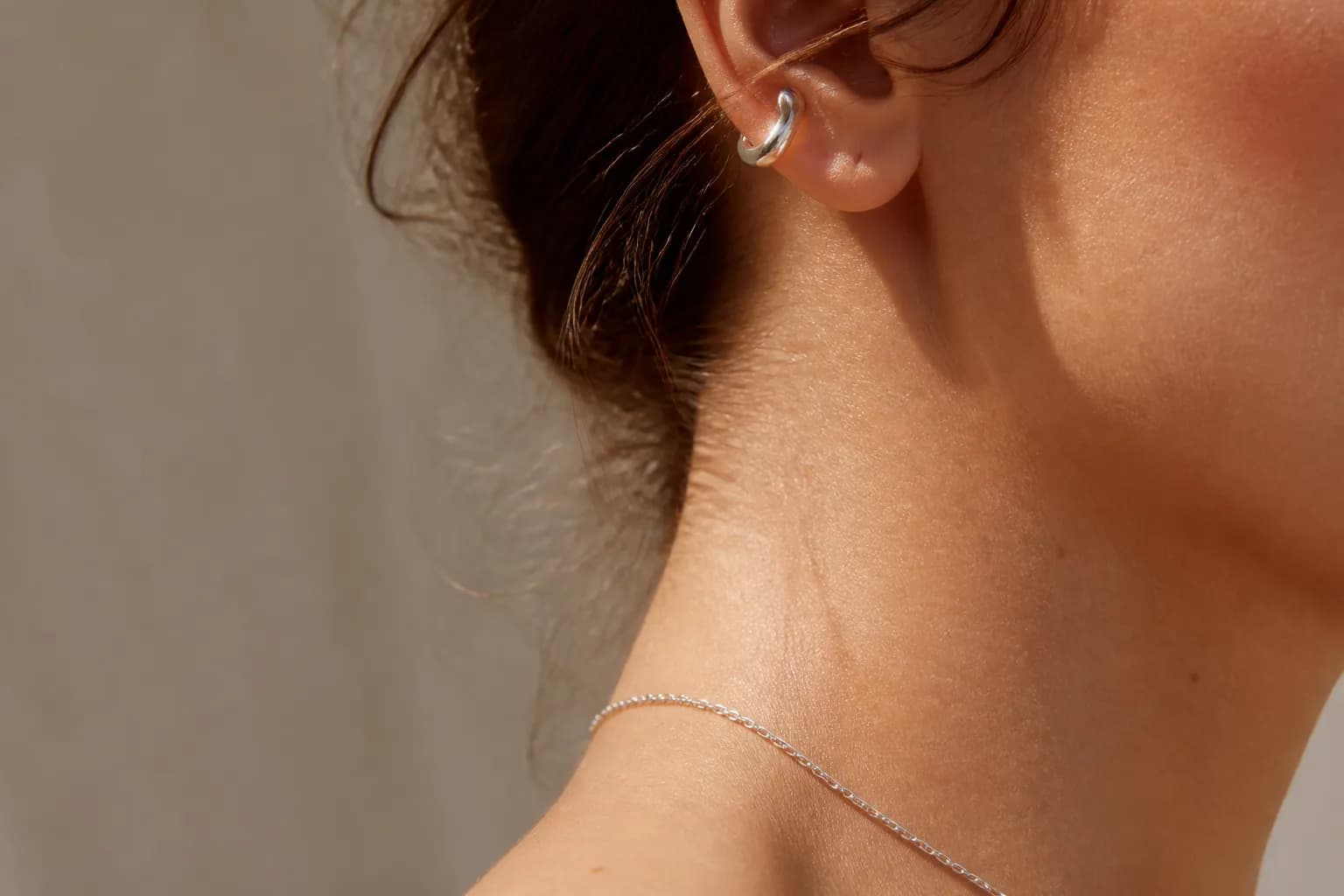 A profile of a neck and ear wearing a small comma-shaped silver ear cuff and a thin chain, chin cropped out of frame.