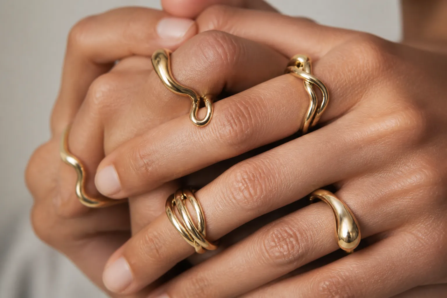 Two interlaced hands wearing several fluid twisted gold rings, knuckles close to the camera against a soft grey-cream backdrop.