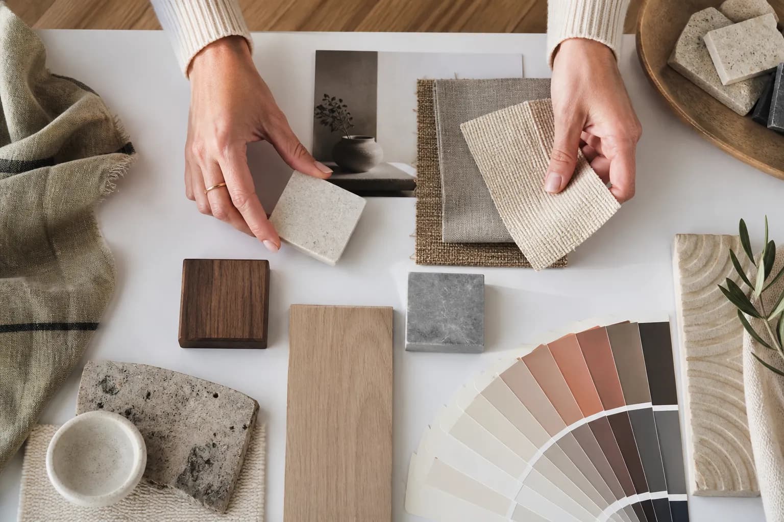 A studio worktable seen from above: hands arranging fabric swatches, wood samples, stone chips and a paint fan on white paper in warm daylight.