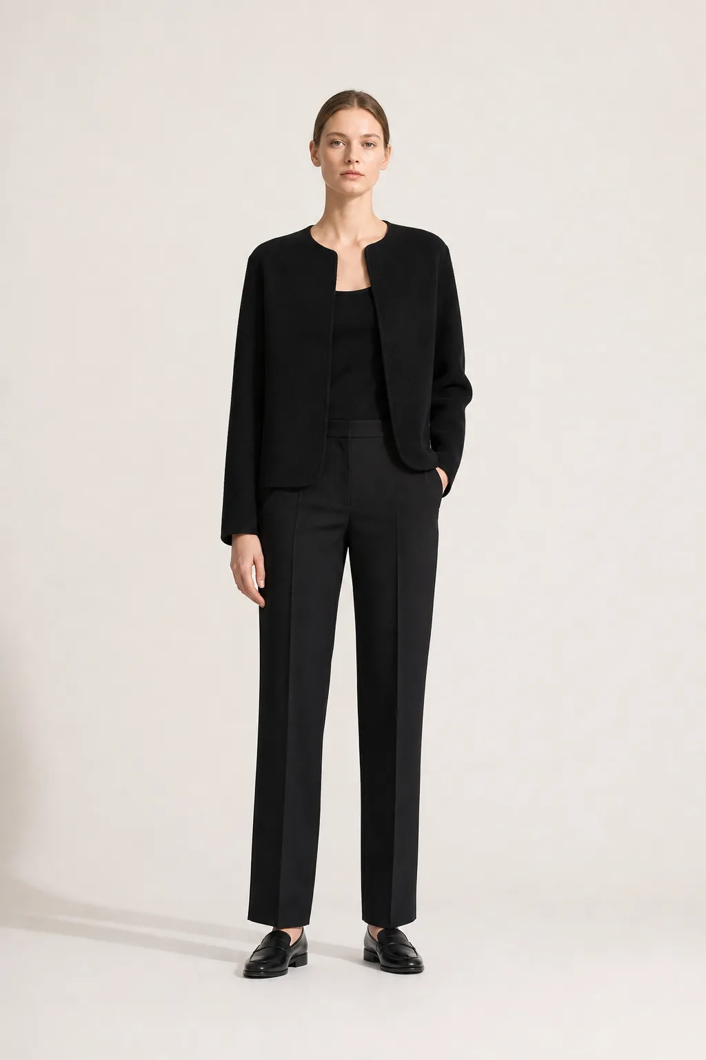 Full-length studio shot of a collarless black wool jacket worn open over straight black trousers and black loafers